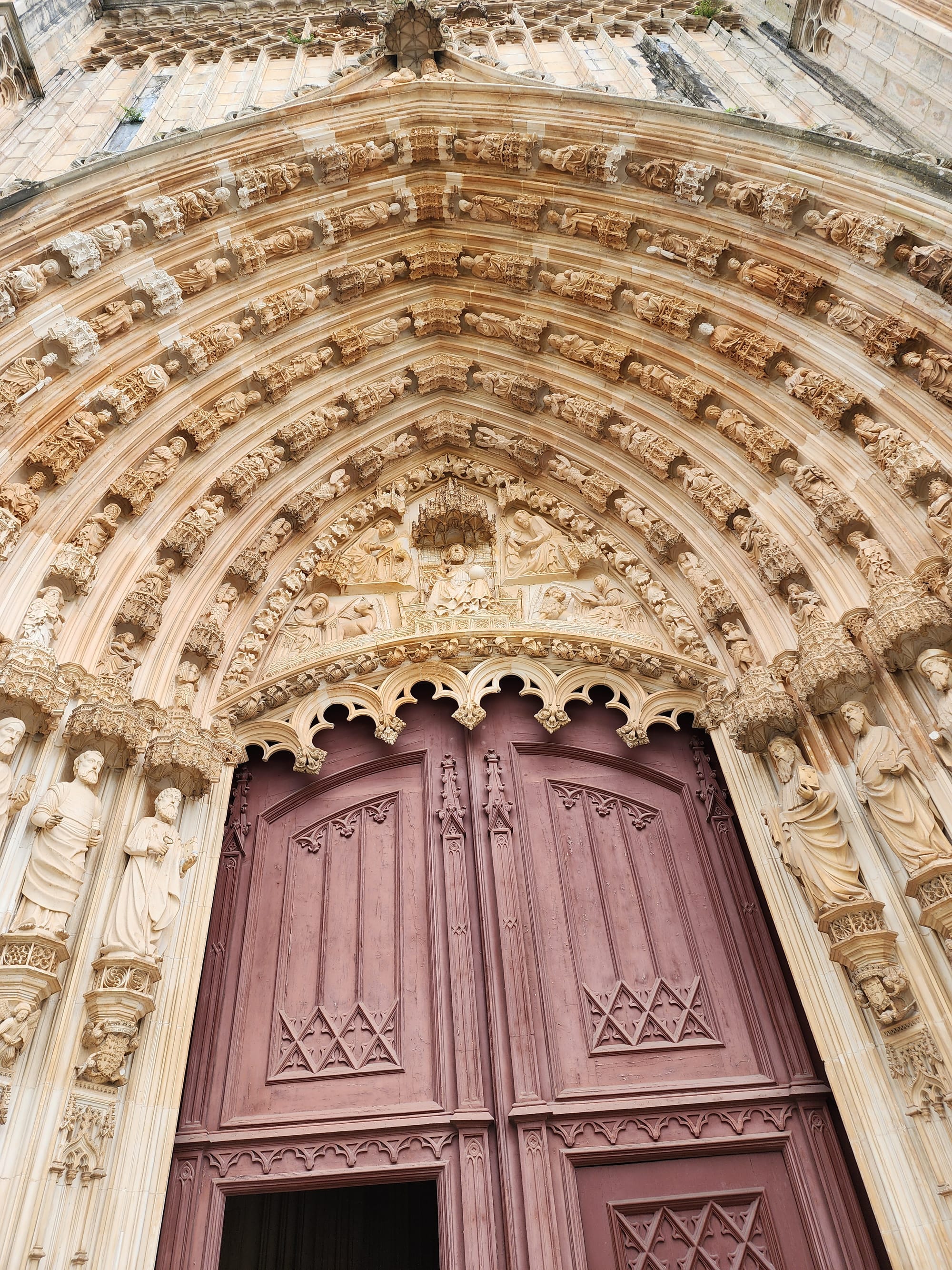 The image showcases a close-up view of a grand, intricately carved Gothic archway leading to large wooden doors. The craftsmanship is detailed with an array of sculpted figures and ornamental motifs, which are characteristic of Gothic ecclesiastical architecture. The rich texture of the stone contrasts with the smooth, weathered surface of the wooden doors, hinting at the countless moments they have witnessed. The historical and artistic significance of such an entrance invites both awe and reverence.